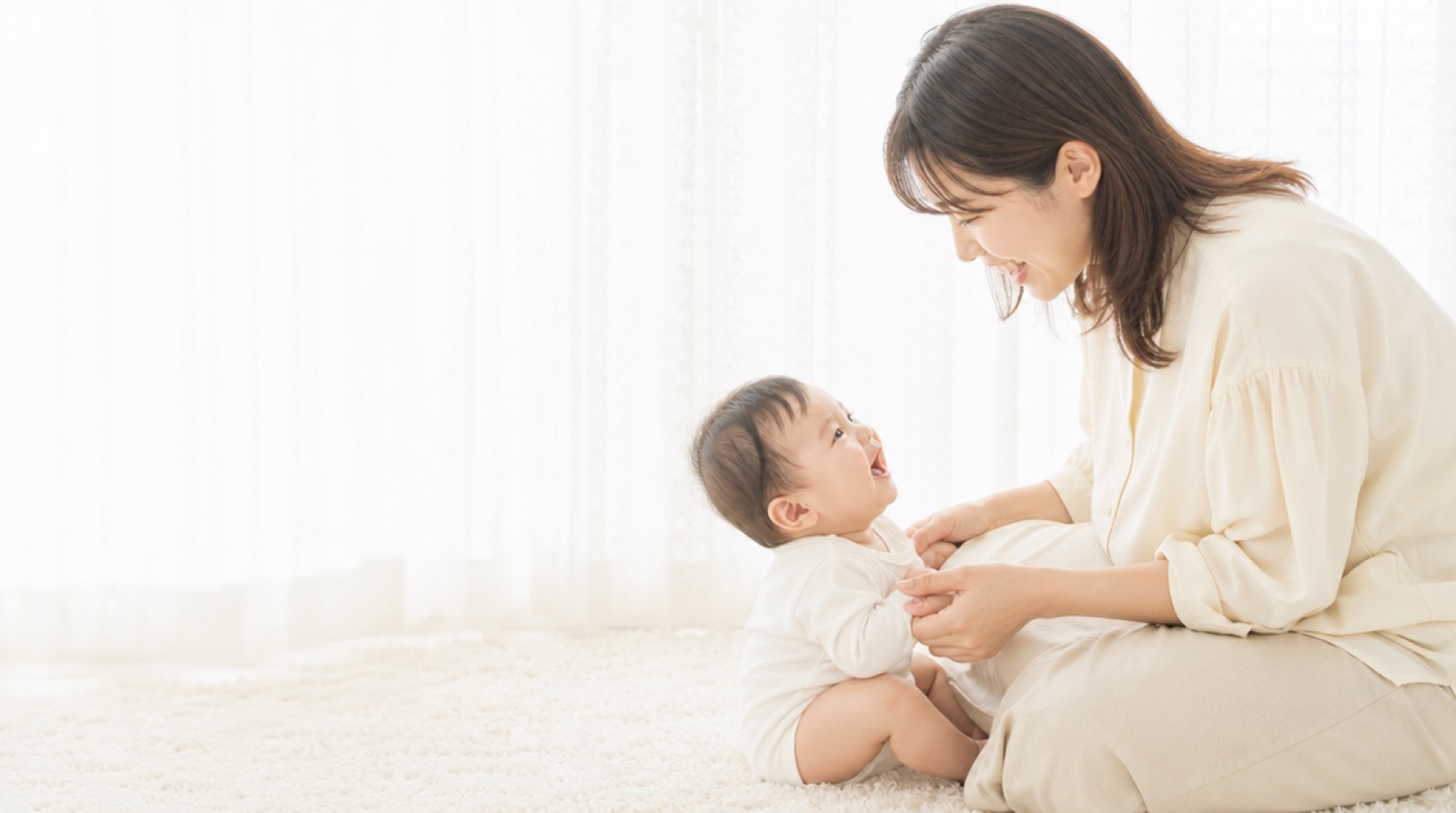 白いラグの上で、母親と赤ちゃんが向かい合って笑い合っている様子