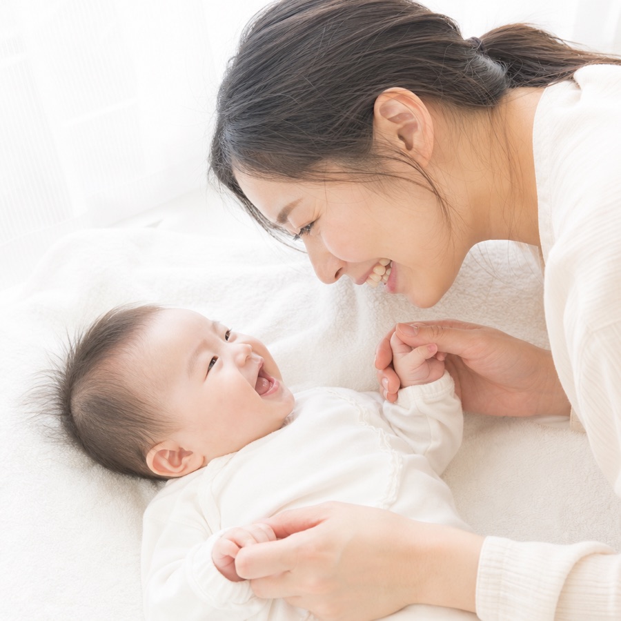 白いタオルの上で笑う赤ちゃんと、顔を近づけて目を合わせる母親